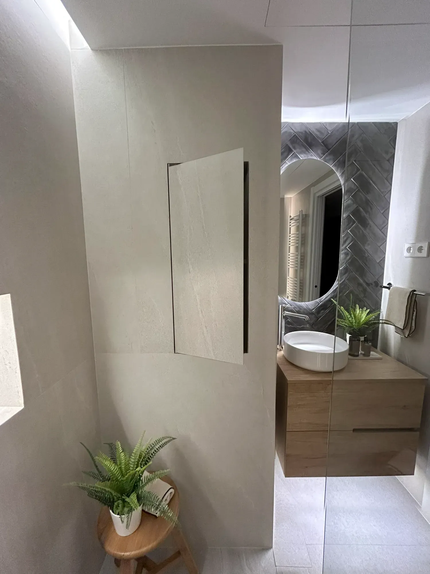Baño moderno con tocador de madera, espejo ovalado y una planta. Paredes blancas y un pequeño taburete.