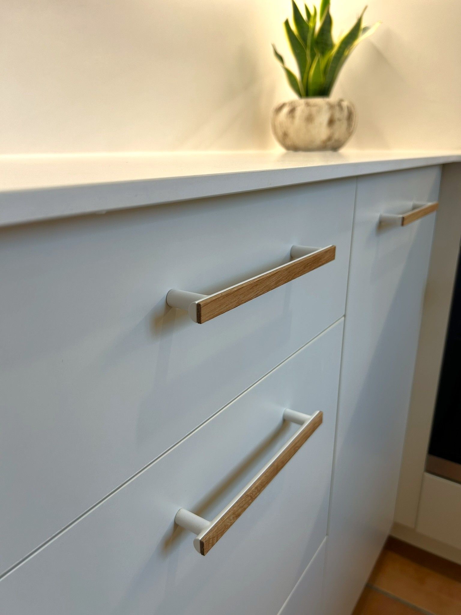Primer plano de cajones de cocina blancos con tiradores modernos de madera y blancos alineados horizontalmente, con una pequeña planta en maceta.
