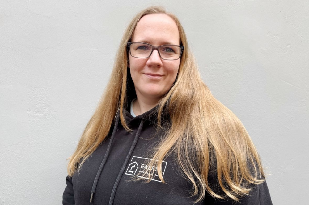 Frau mit langen blonden Haaren und Brille, trägt einen schwarzen Kapuzenpullover und steht vor einer grauen Wand.