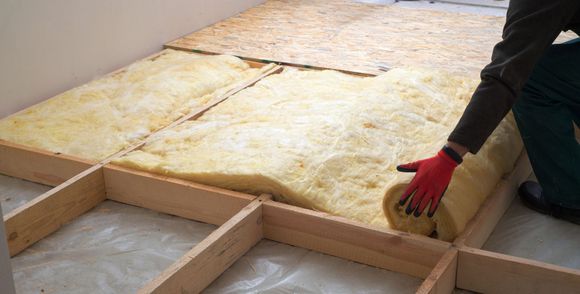 Une personne portant des gants rouges installe des panneaux isolants entre les solives en bois d'un plancher à l'intérieur d'une pièce.