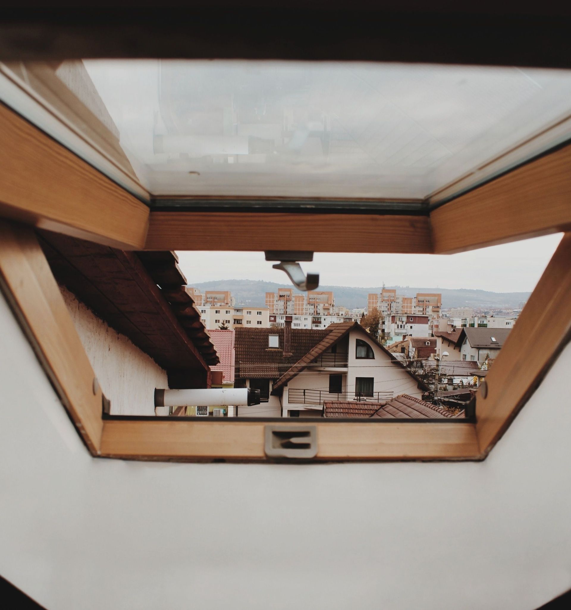 Vue à travers une lucarne ouverte sur des maisons aux toits rouges et la silhouette de la ville sous un ciel nuageux