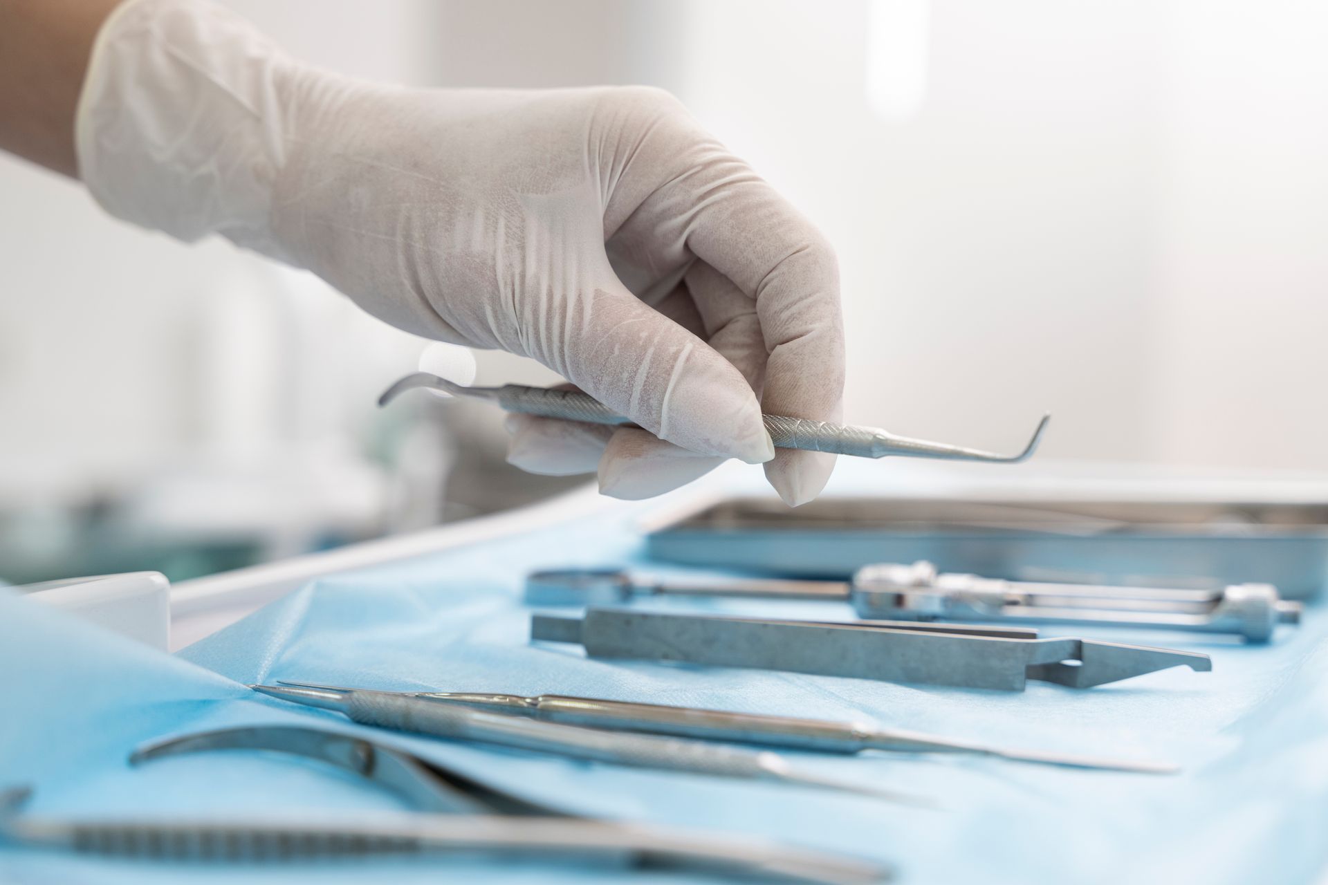Mano enguantada sosteniendo un instrumento dental sobre una bandeja de instrumentos de acero inoxidable en una clínica.