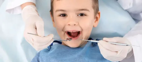 Un niño sentado en un sillón dental con la boca abierta mientras un dentista le examina los dientes con instrumentos y guantes.