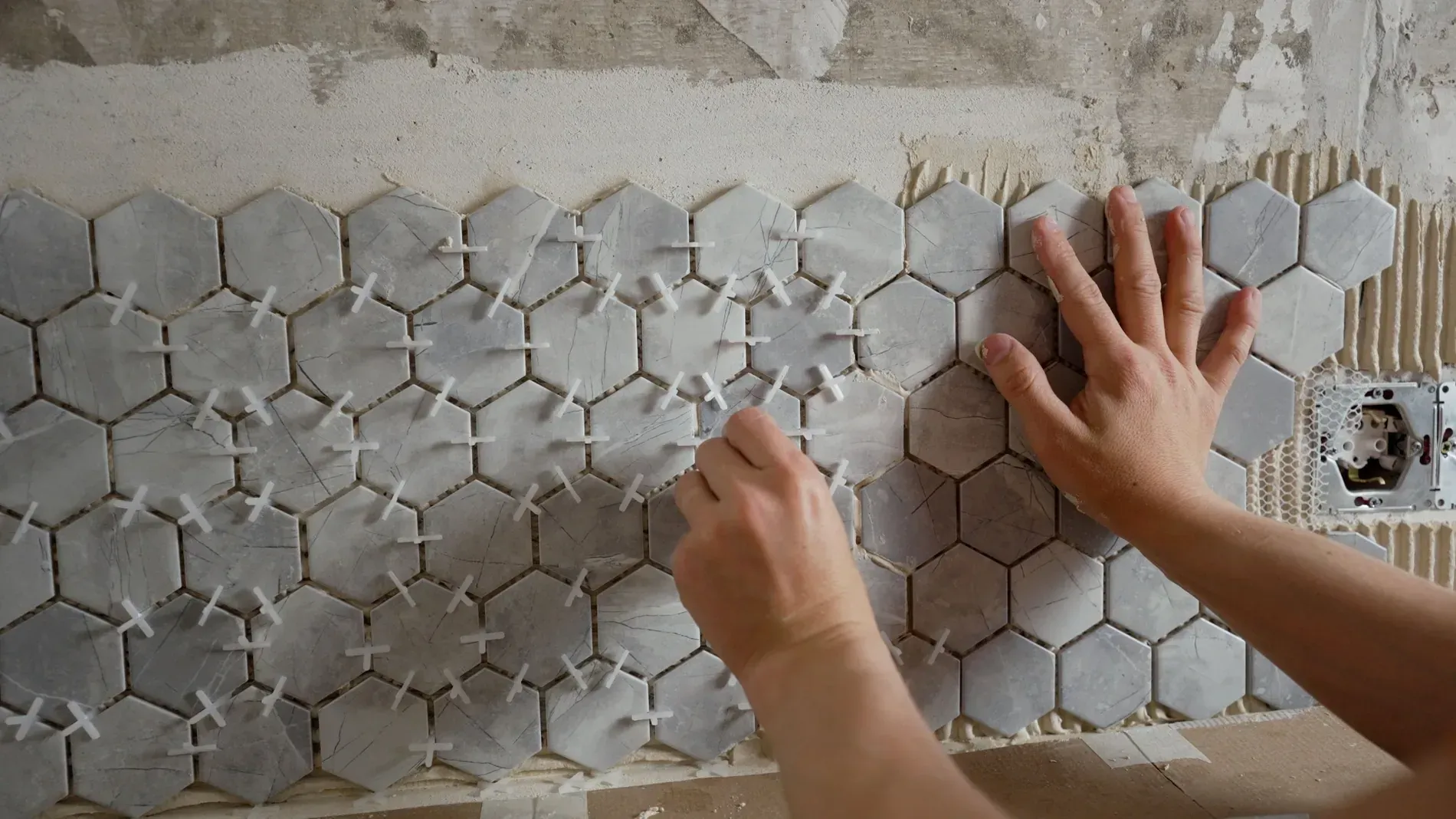 Manos colocando baldosas hexagonales con espaciadores en una pared, construcción en progreso.