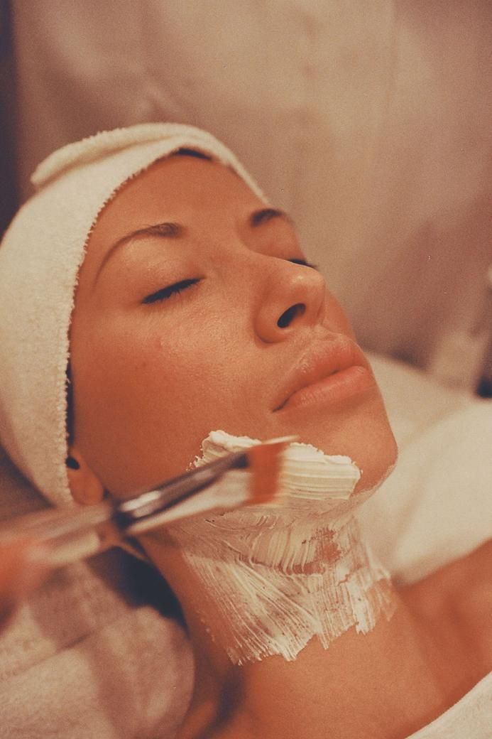 Persona a la que se le aplica una mascarilla facial con un pincel, tumbada boca arriba con una diadema y los ojos cerrados.