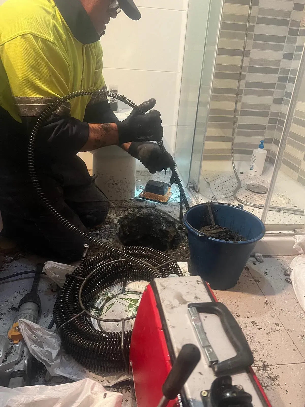 Un hombre está arrodillado en el suelo de un baño usando un limpiador de desagües.