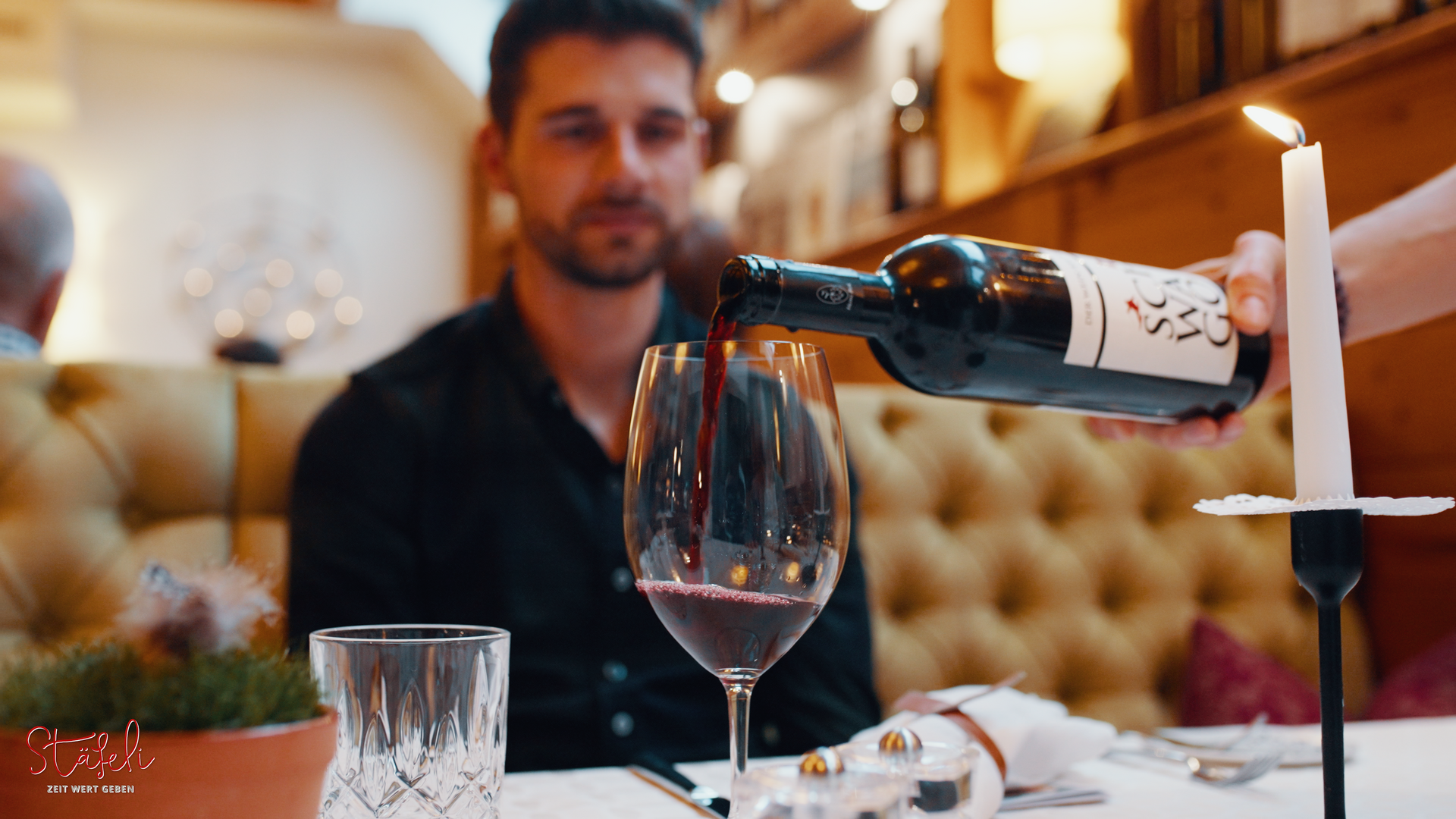 Ein Mann sitzt an einem Tisch und es wird Wein aus einer Flasche eingeschenkt.