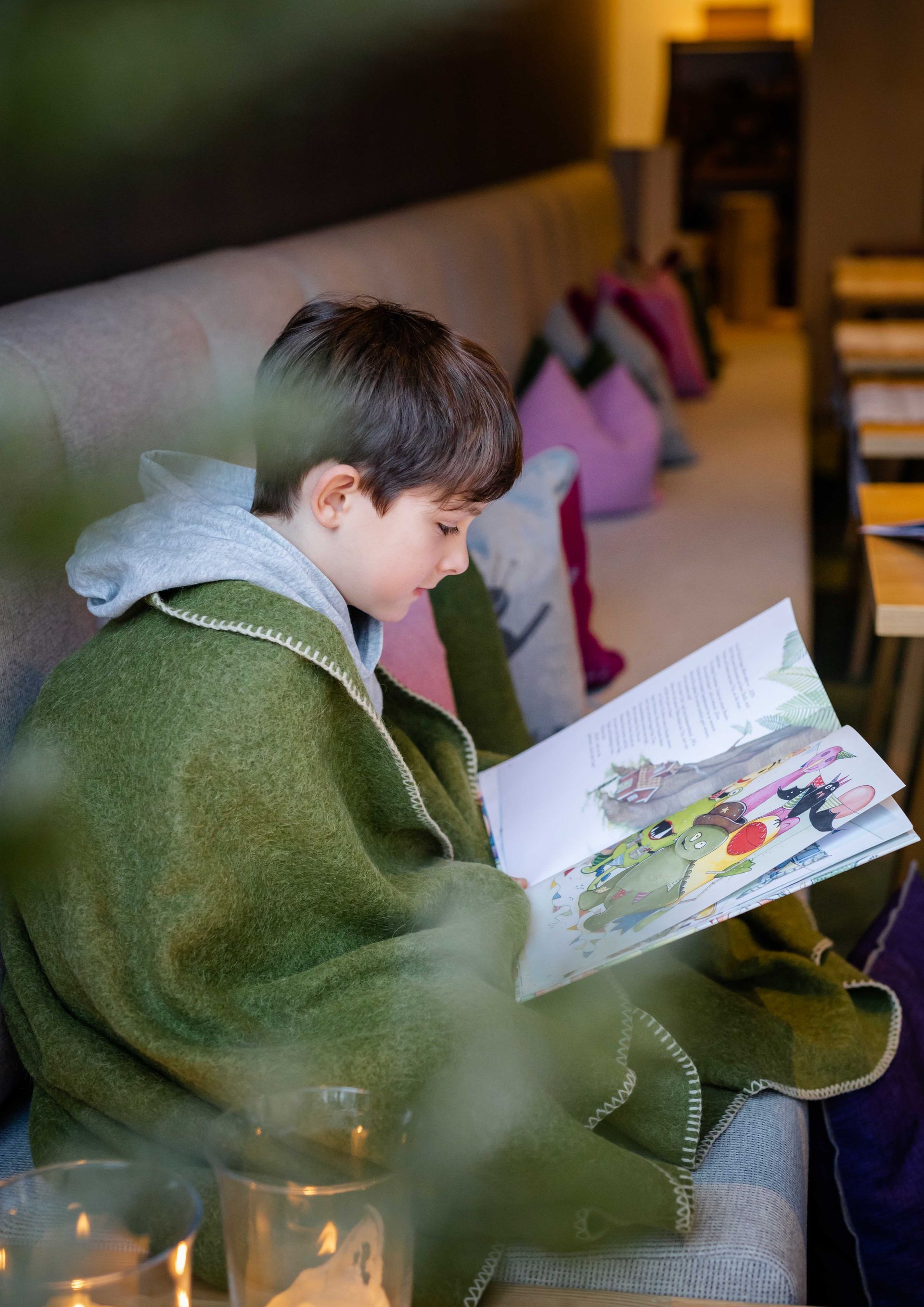 Ein Junge , der mit einer grünen Decke bedeckt ist , sitzt auf einer Couch und liest ein Buch .