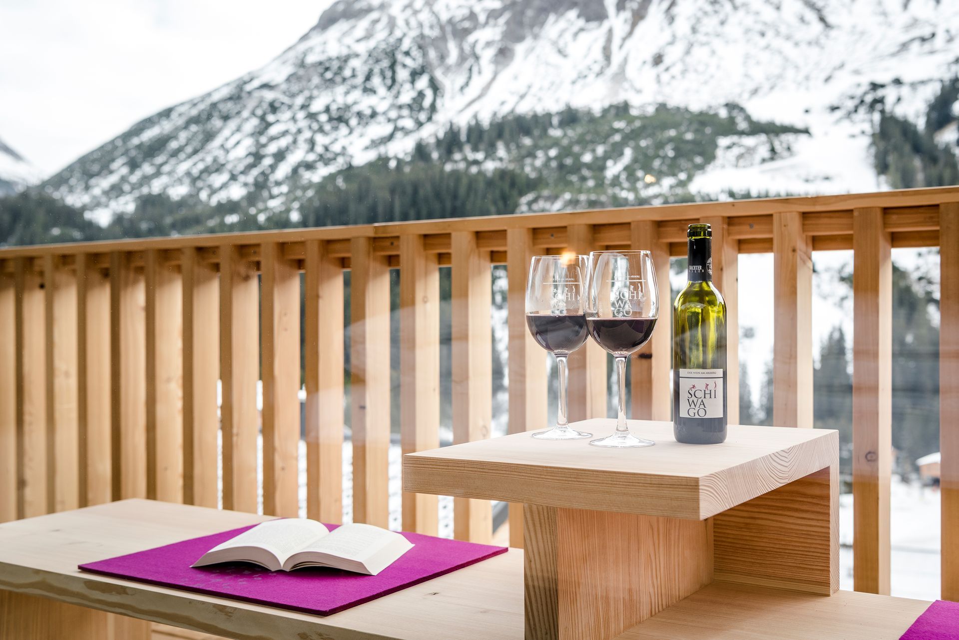 Auf einem Holztisch auf dem Balkon stehen zwei Gläser Wein und eine Flasche Wein vor winterlicher Bergkulisse .