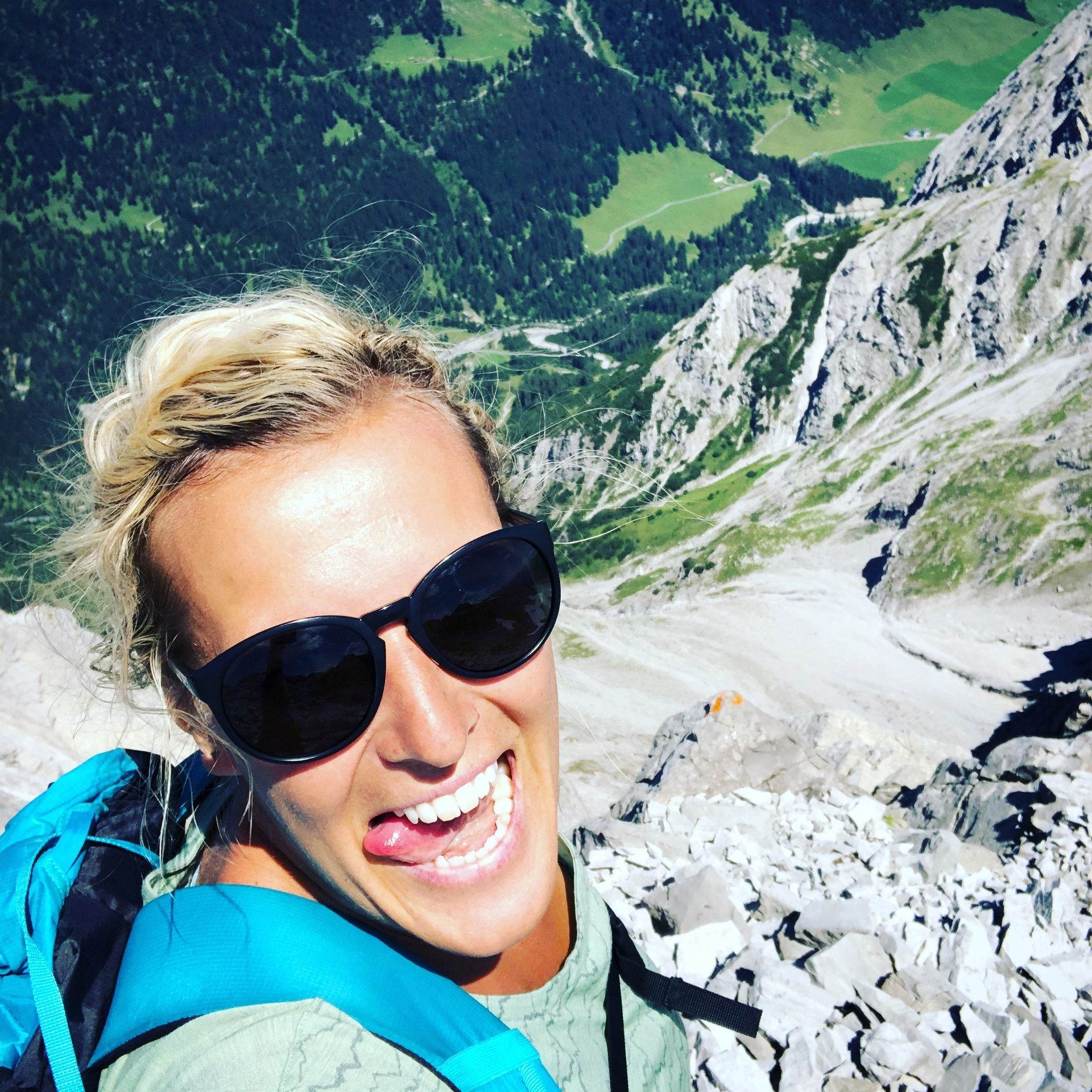 Eine Frau mit Sonnenbrille und Rucksack lächelt auf einem Berg