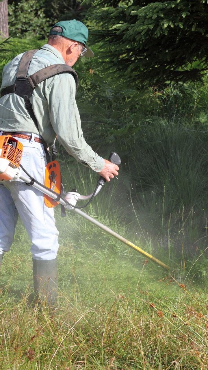 Une personne en chemise et chapeau verts utilise une débroussailleuse dans un champ herbeux.