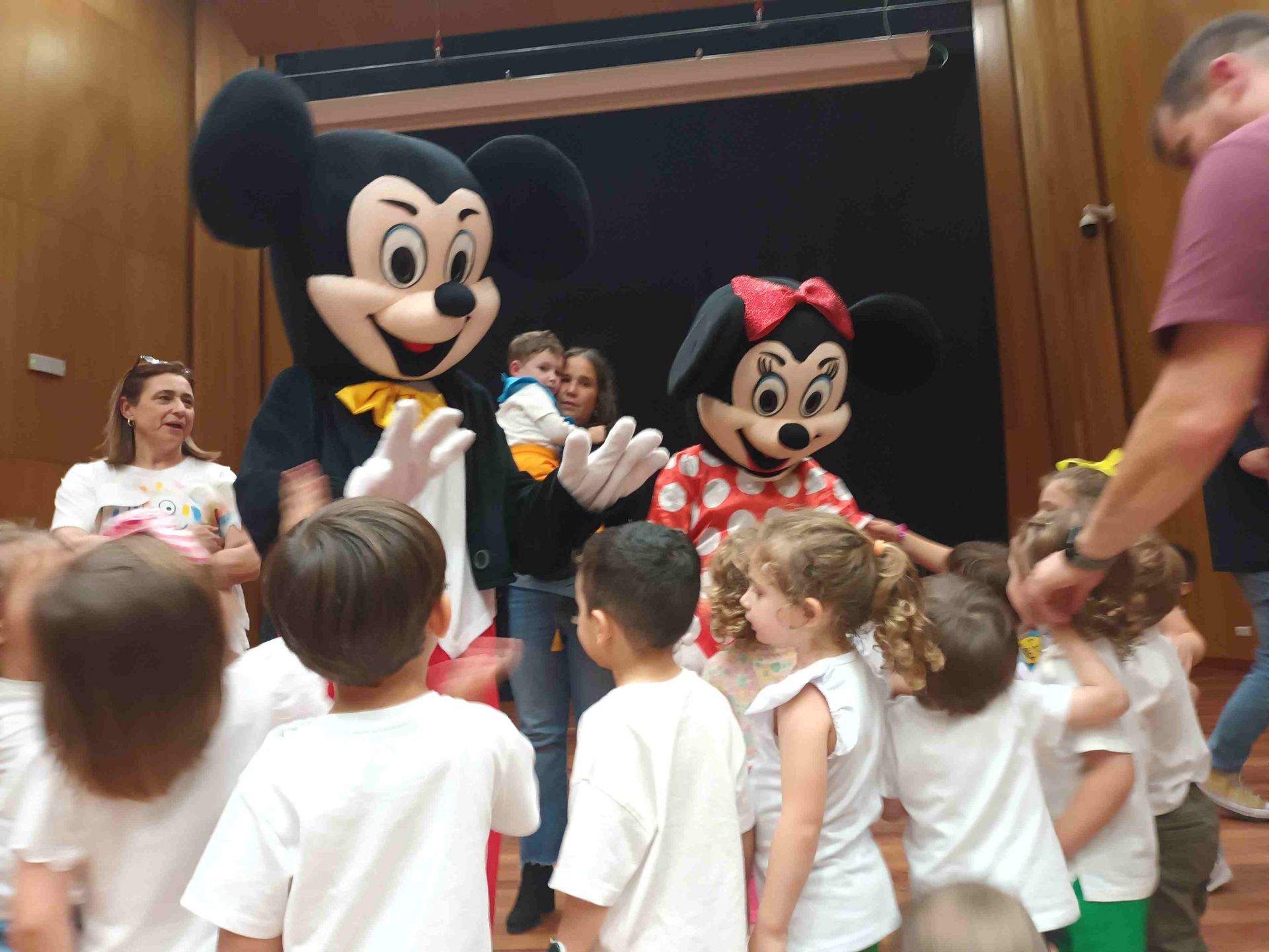 Mickey y Minnie Mouse con niños frente a un escenario. Hay gente presente.