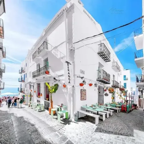 Cafetería de un blanco brillante en una calle empedrada bajo un cielo azul, con mesas, plantas en macetas y flores colgantes.