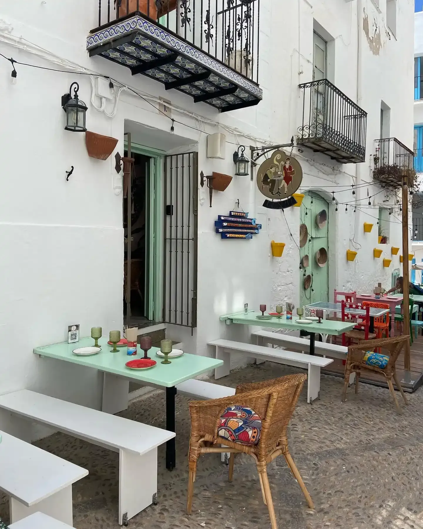 Mesas y sillas de café al aire libre junto a edificios blancos con balcones en una estrecha calle empedrada.