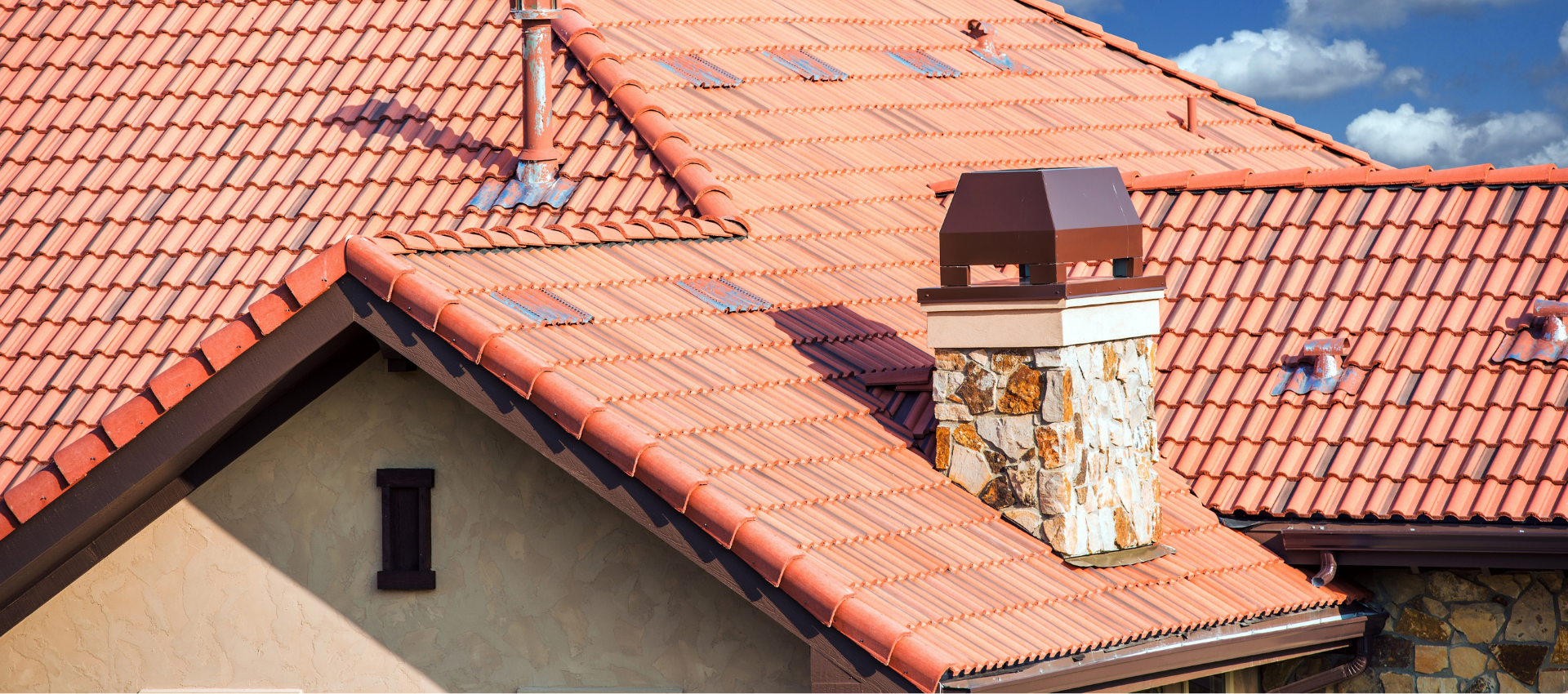 Toit de tuiles rouges, cheminée en pierre et boiseries brunes se détachant sur un ciel bleu.