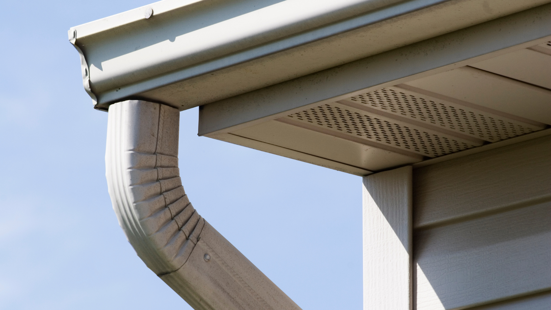 Coin de maison beige avec gouttière et descente pluviale, sur fond de ciel bleu.