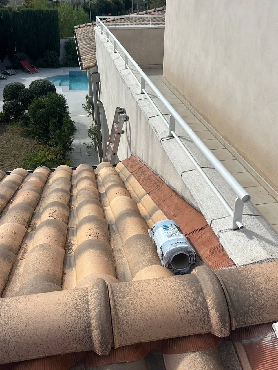 Toiture avec tuiles orange et rambarde de sécurité blanche, un outil de mesure laser et une échelle adossée au mur.
