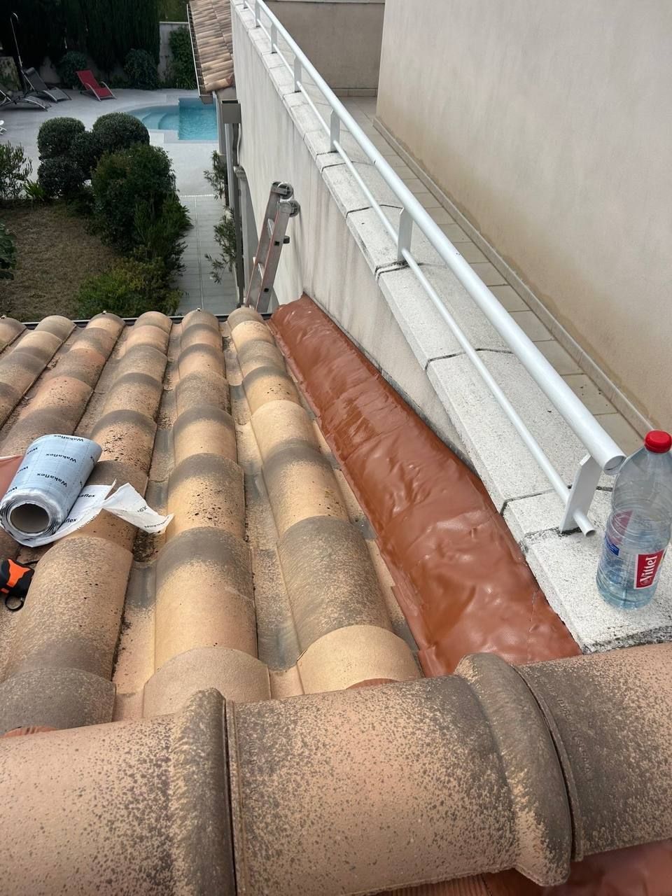 Toiture en tuiles de terre cuite, gouttière et rambarde blanche. Les matériaux de construction sont visibles.