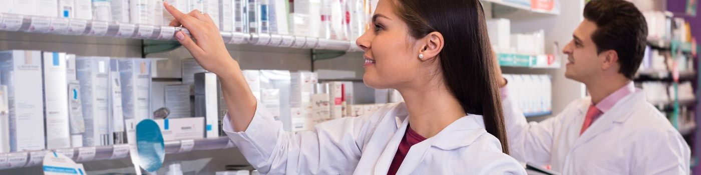 Dos farmacéuticos con batas blancas alcanzan los productos de un estante de la farmacia, sonriendo.