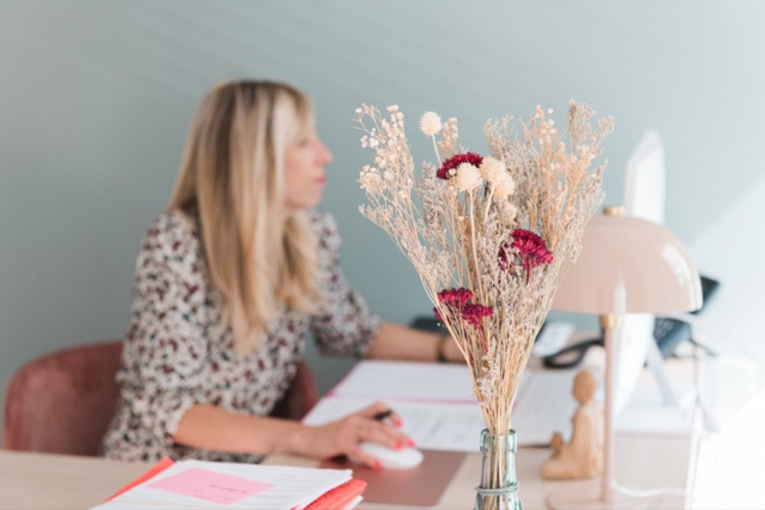 Fleurs séchées sur un bureau ; femme floue en arrière-plan travaillant sur un ordinateur.