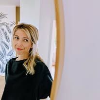 Une femme en haut noir sourit en regardant un miroir rond. Cheveux blonds, intérieur.