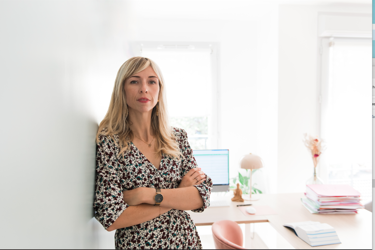 Femme en robe à fleurs, les bras croisés, appuyée contre un mur blanc dans un bureau avec un bureau.