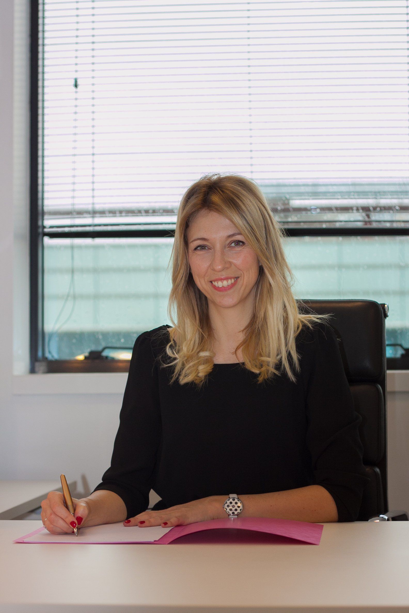 Femme en robe noire souriante, écrivant à un bureau, dossier rose, cadre de bureau moderne.