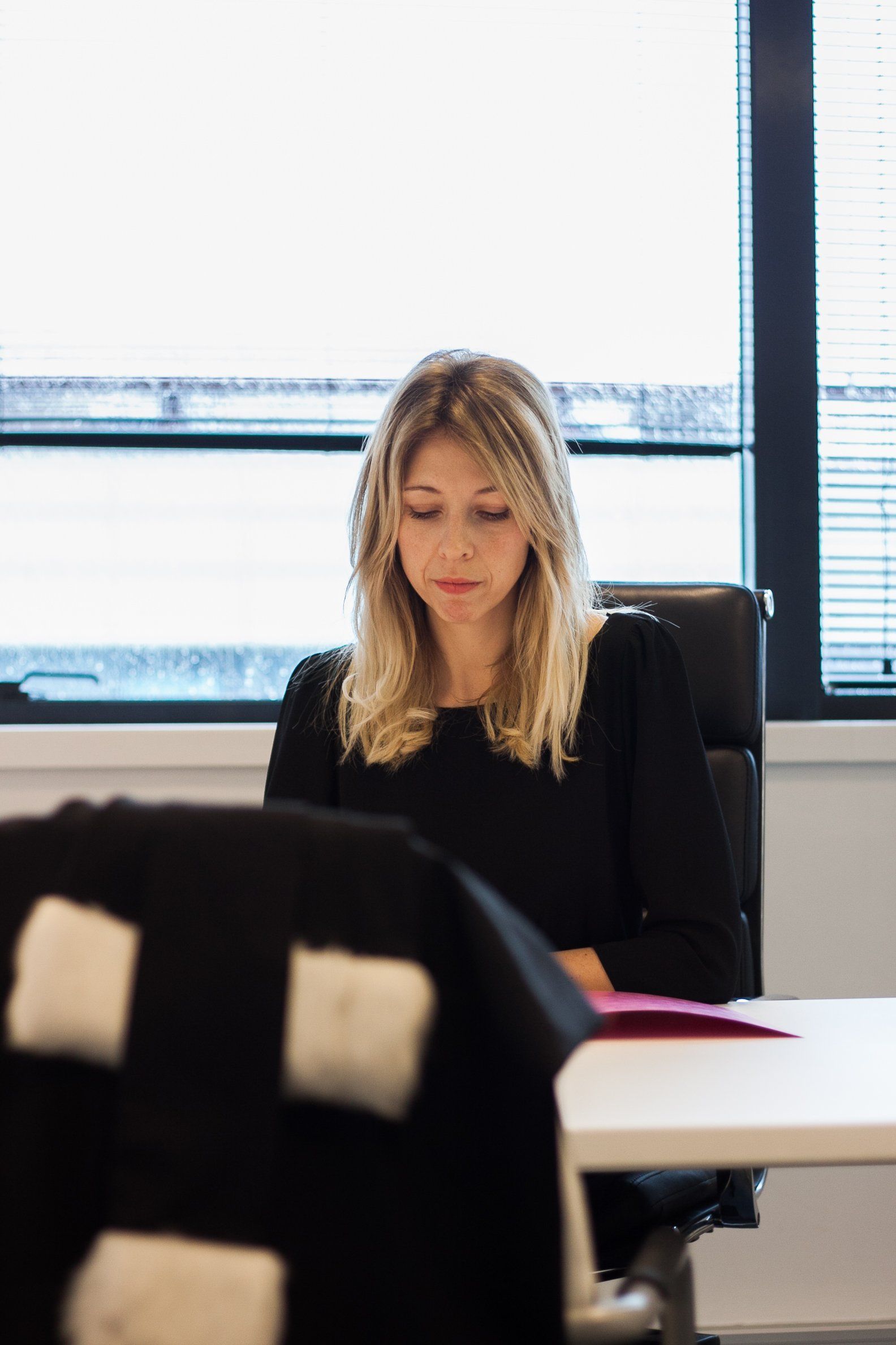 Une femme en robe noire est assise à un bureau, le regard baissé. Stores et fenêtre en arrière-plan.