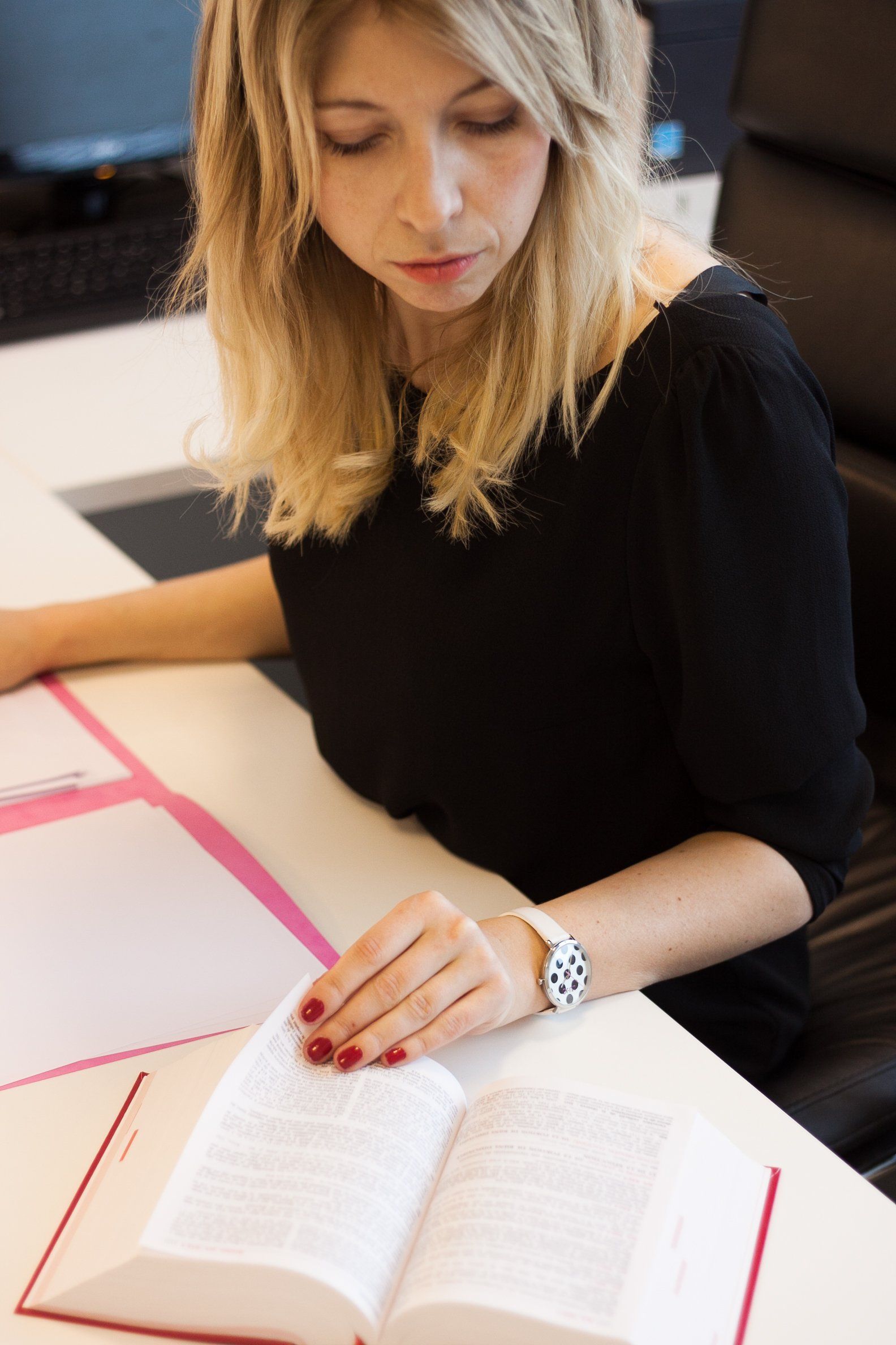 Femme blonde lisant un livre à une table blanche, vêtue d'un haut noir et portant une montre.