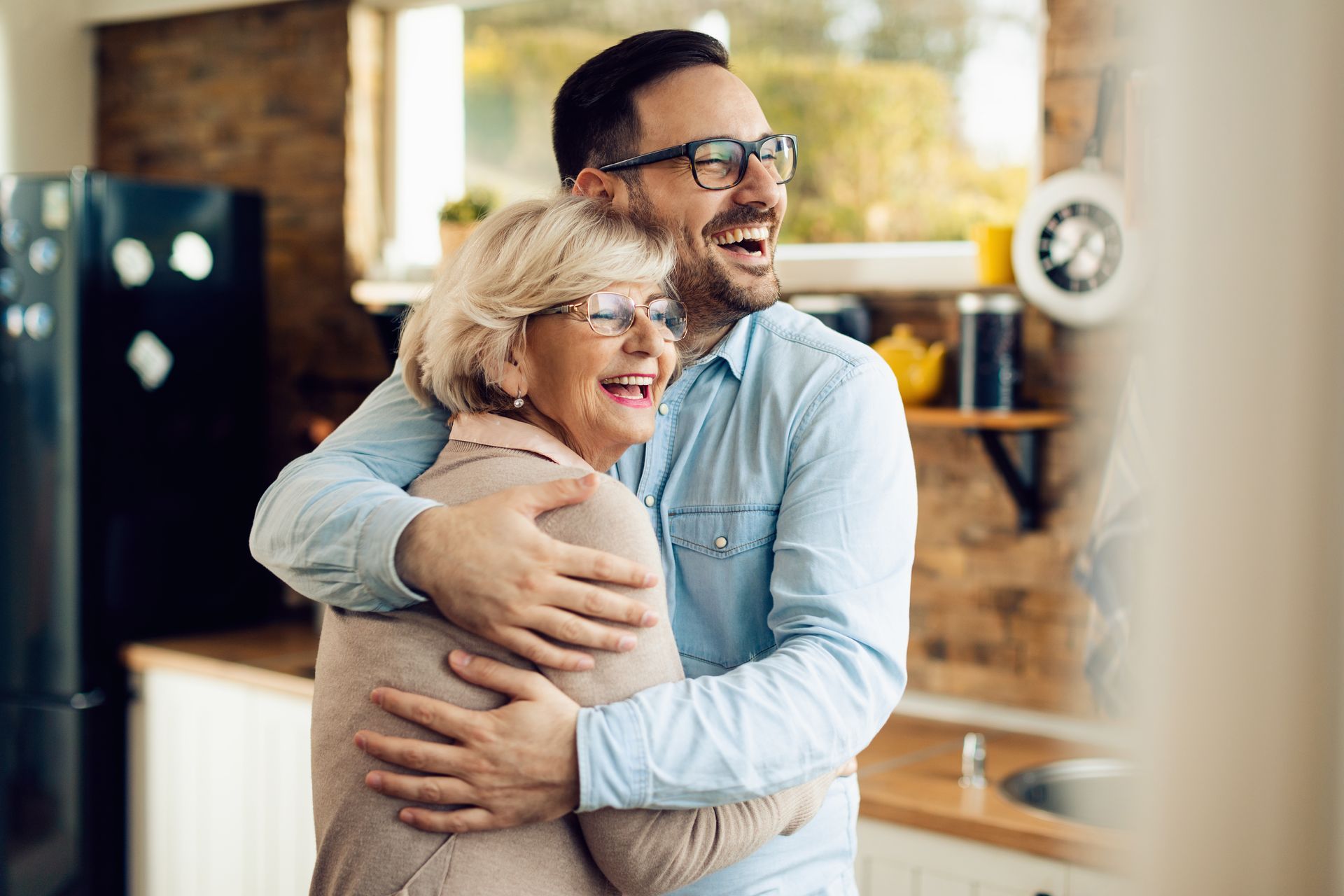 Un homme enlace une femme, tous deux souriants dans une cuisine. La lumière du soleil inonde la pièce.