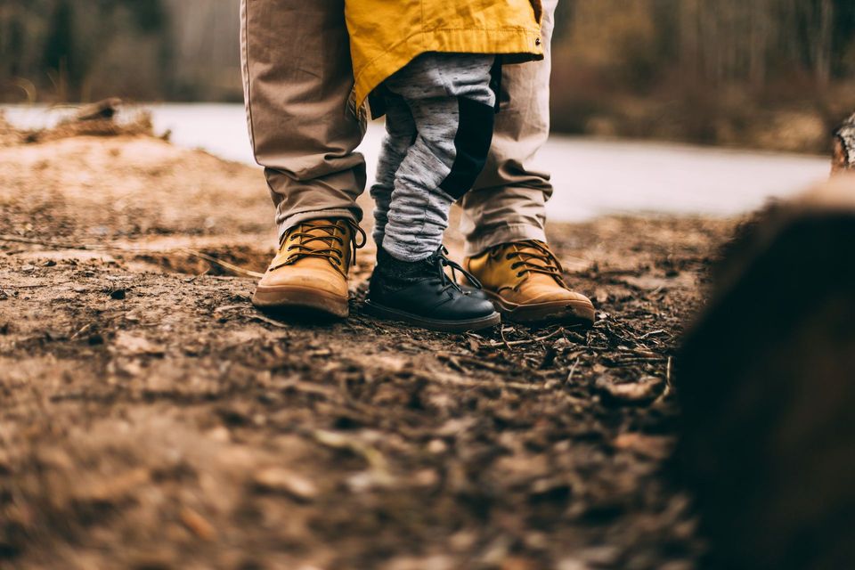 Trois paires de pieds bottés, posés sur la terre : des jambes d'adulte en pantalon beige à côté de jambes d'enfant en pantalon gris et noir.