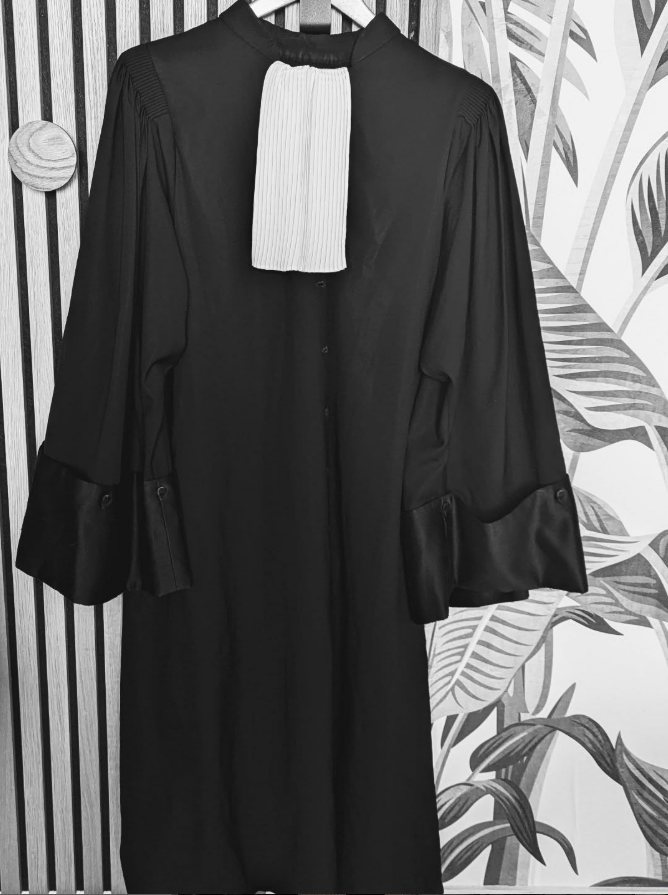 Robe noire d'avocat, à jabot blanc suspendue à un cintre, sur un papier peint à rayures et à motifs floraux.