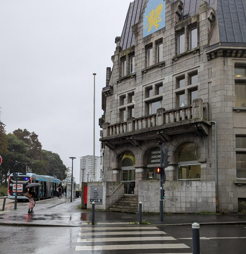 Extérieur d'un bâtiment en pierre avec balcon, passage piéton, tramway et ciel couvert.