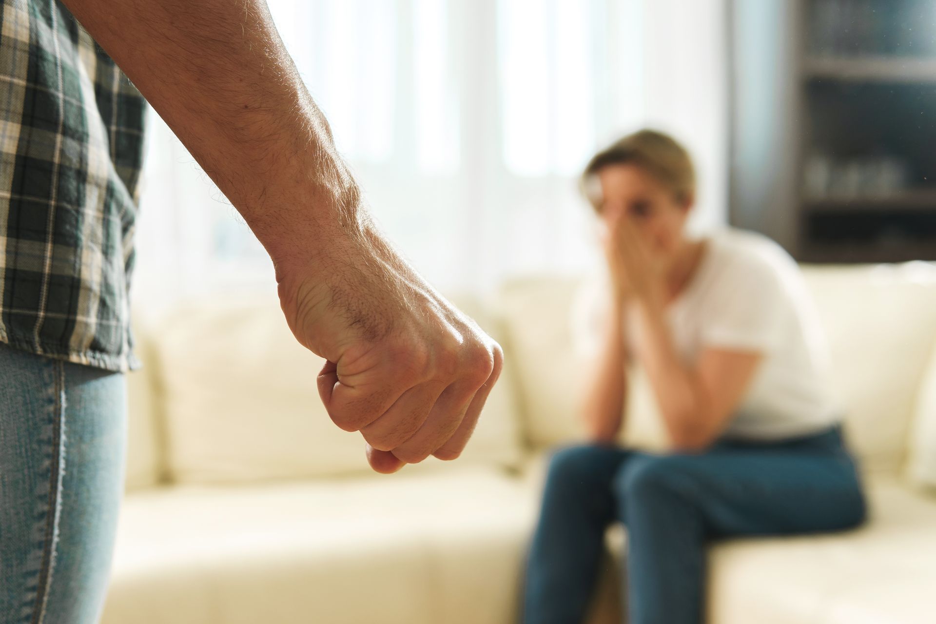 Un homme lève le poing, une femme en arrière-plan se couvre le visage. Conflit représenté dans un cadre domestique.