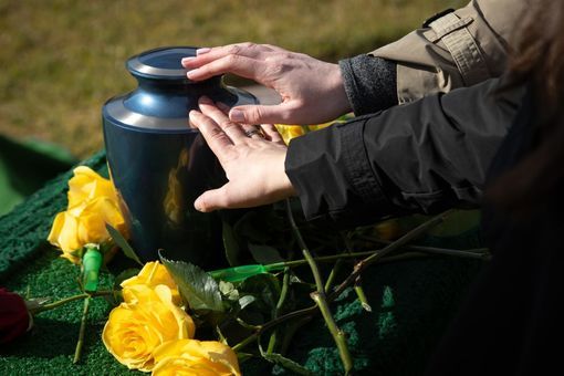 Zwei Personen legen ihre Hände auf eine dunkle Urne, die von gelben Rosen umgeben ist