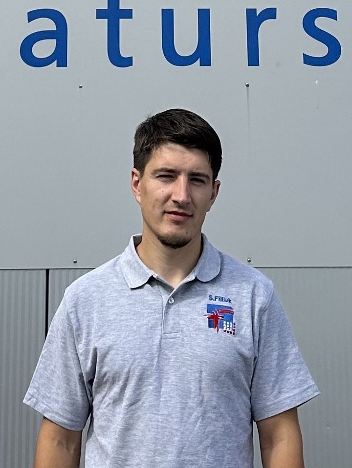 Ein Mann in einem grauen Poloshirt steht vor einem Gebäude mit blauer Schrift.