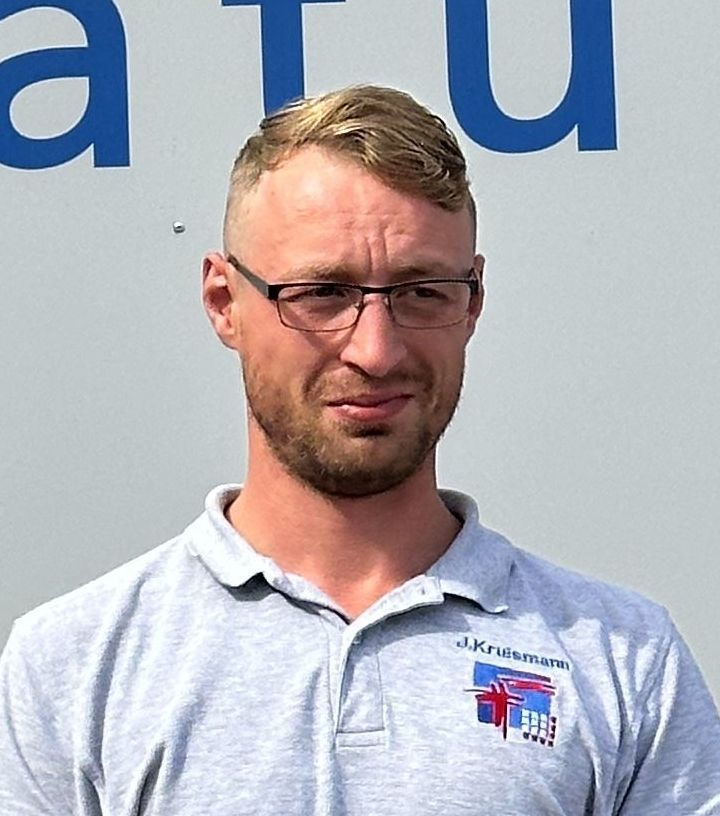 Ein Mann mit Brille und kurzem Haar, der ein graues Poloshirt trägt, steht vor einer weißen Fläche.