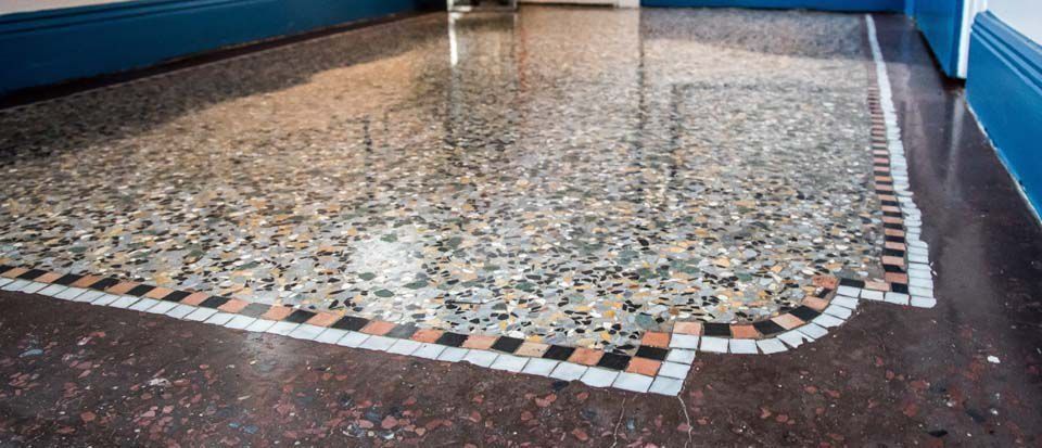 Terrazzoboden mit gemusterter Bordüre, der das Licht reflektiert. Blaue Zierleiste.