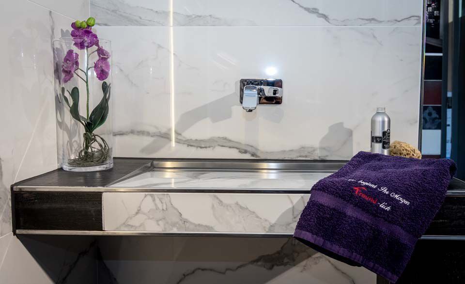 Waschbecken im Badezimmer mit Marmorfliesen, lila Handtuch, Blume in einer Vase und Seifenspender.