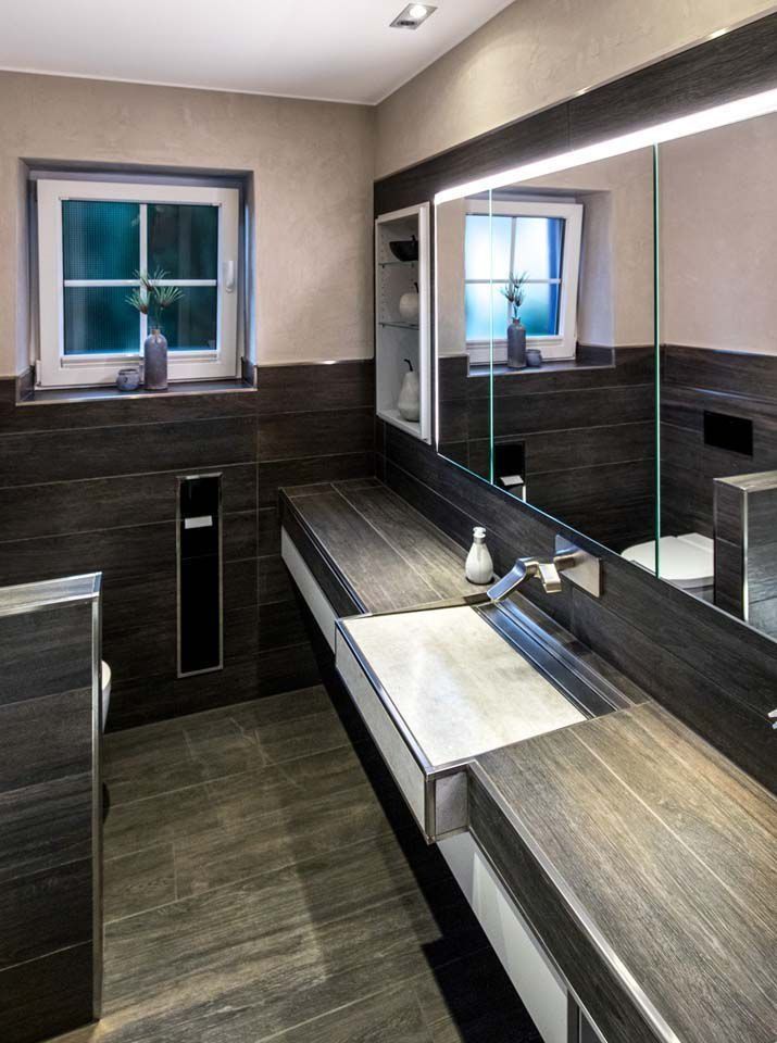 Modernes Badezimmer mit dunklen Holzmaserungsflächen, einem langen Waschbecken und einem großen Spiegel mit integrierter Beleuchtung.