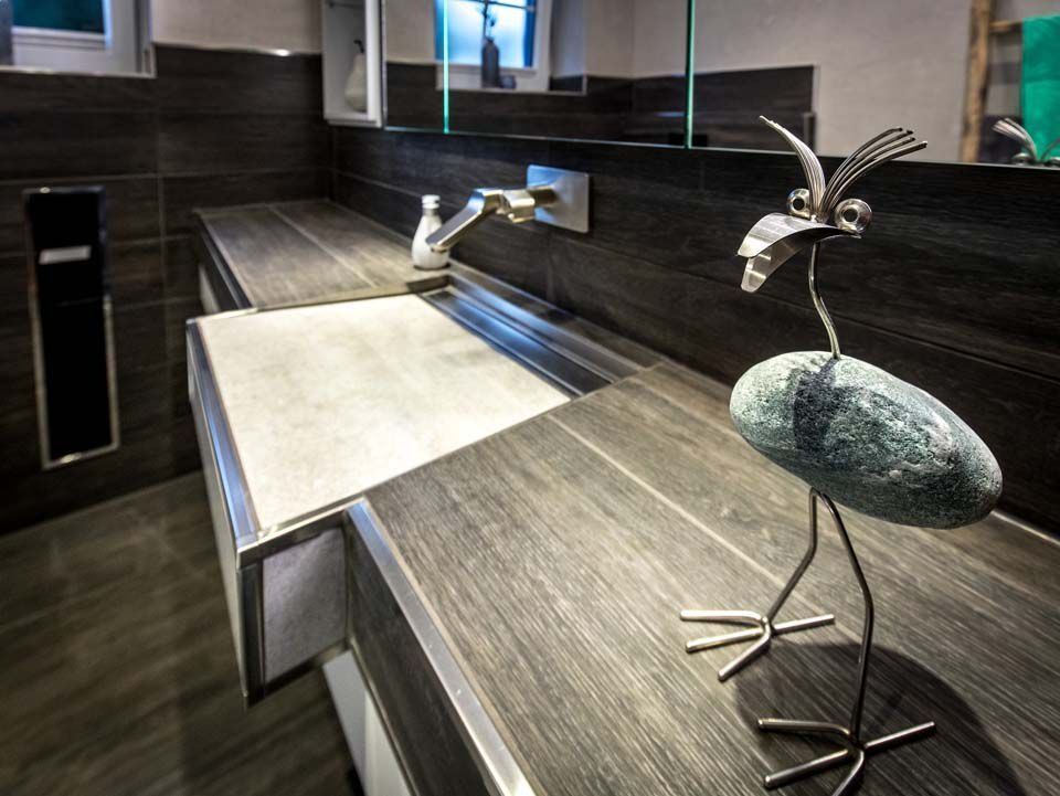 Modernes Badezimmer mit dunkler Holzmaserung auf der Arbeitsplatte, integriertem Waschbecken und Vogelskulptur aus Metall.