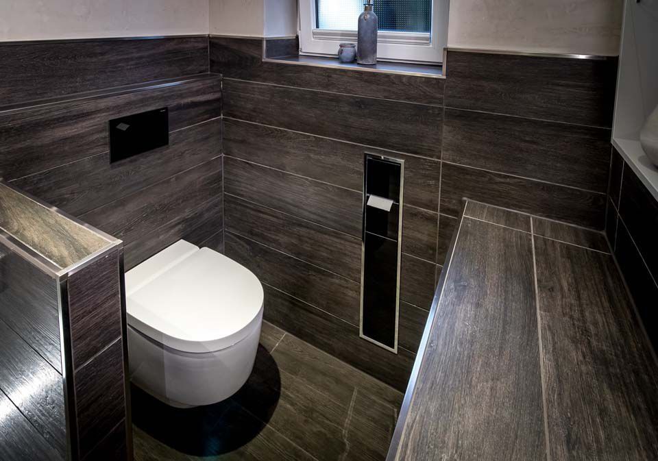 Modernes Badezimmer mit dunklen Fliesen in Holzoptik. Mittig angeordnetes WC mit Spülkasten. Kleines Fenster mit Dekoration.