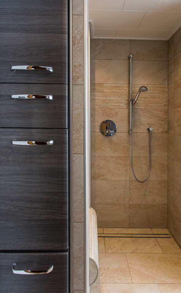 Ein dunkler Holzschrank neben einer gefliesten Duschkabine. Armaturen aus Chrom, neutrale beige Fliesen und ein weißes Handtuch.