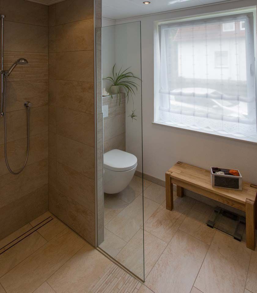 Badezimmer mit verglaster Dusche, Toilette, Holzbank und Fenster mit transparentem Vorhang.