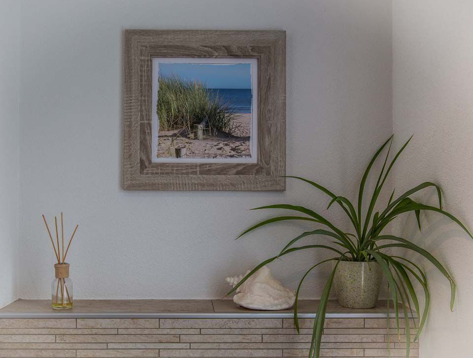 Fotografie einer Küstenlandschaft in einem rustikalen Holzrahmen, über einem Regal mit einer Muschel, einem Diffusor und einer Topfpflanze.