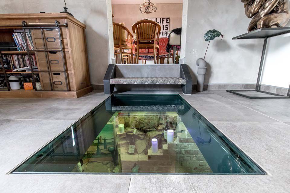 Eine mit Glas abgedeckte Grube im Boden gibt den Blick auf einen unterirdischen Raum mit Möbeln und Dekoration frei.