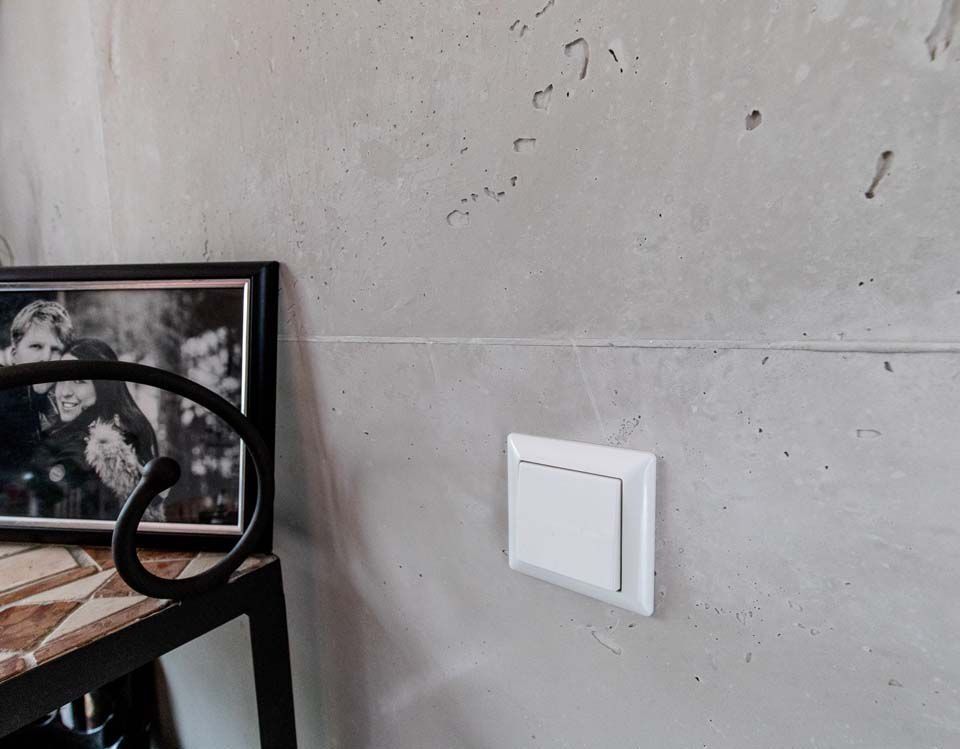 Ein weißer Lichtschalter an einer strukturierten grauen Betonwand neben einem gerahmten Schwarzweißfoto.