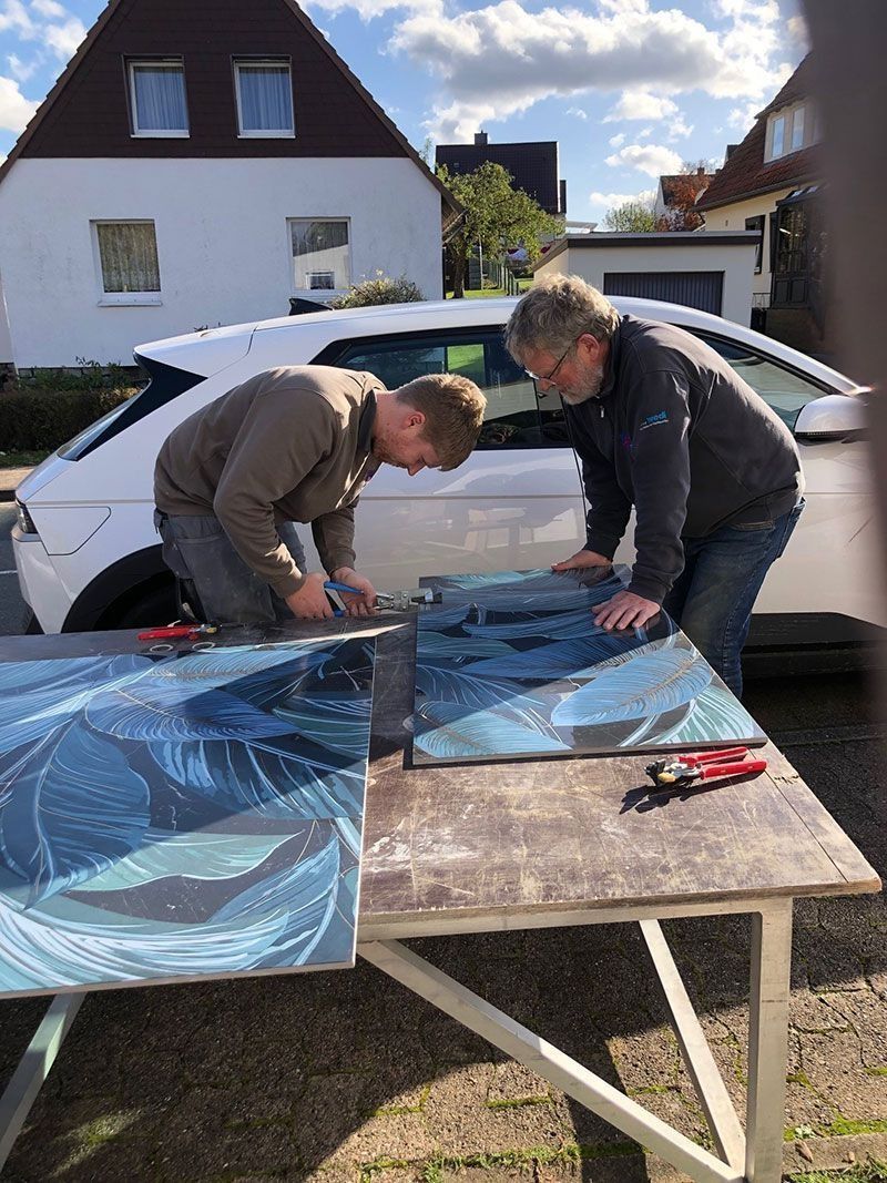 Zwei Personen schneiden Material an einem Tisch im Freien zu; im Hintergrund sind ein weißes Auto und ein Haus zu sehen.