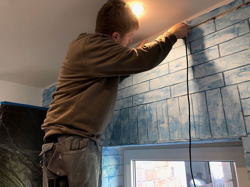Eine Person montiert Zierleisten an einer Wand, die so gestrichen ist, dass sie blaue Ziegelsteine ​​darstellt, in der Nähe eines Fensters.