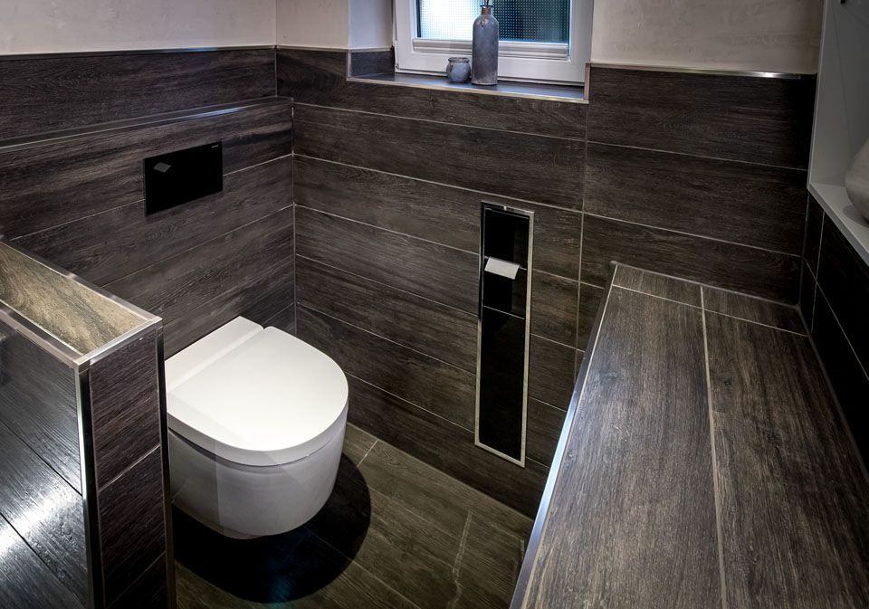 Dunkel gefliestes Badezimmer mit wandhängender Toilette, Toilettenpapierspender und kleinem Fenster.